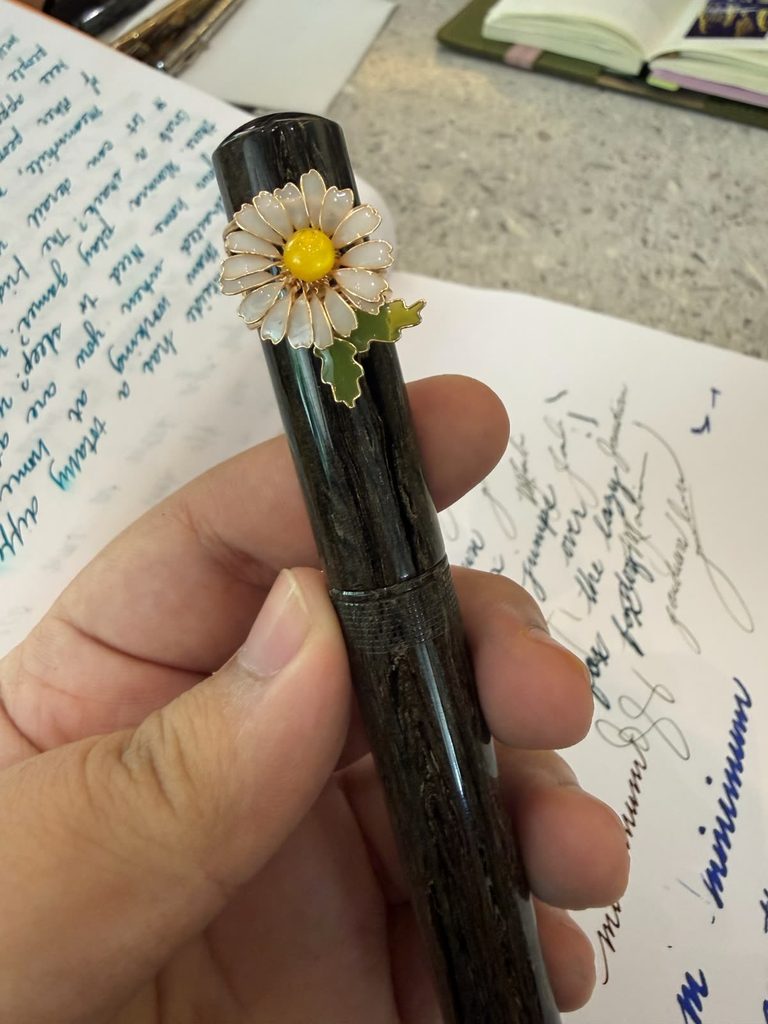A vintage fountain pen with a daisy-shaped cap decoration, held over handwritten journal pages